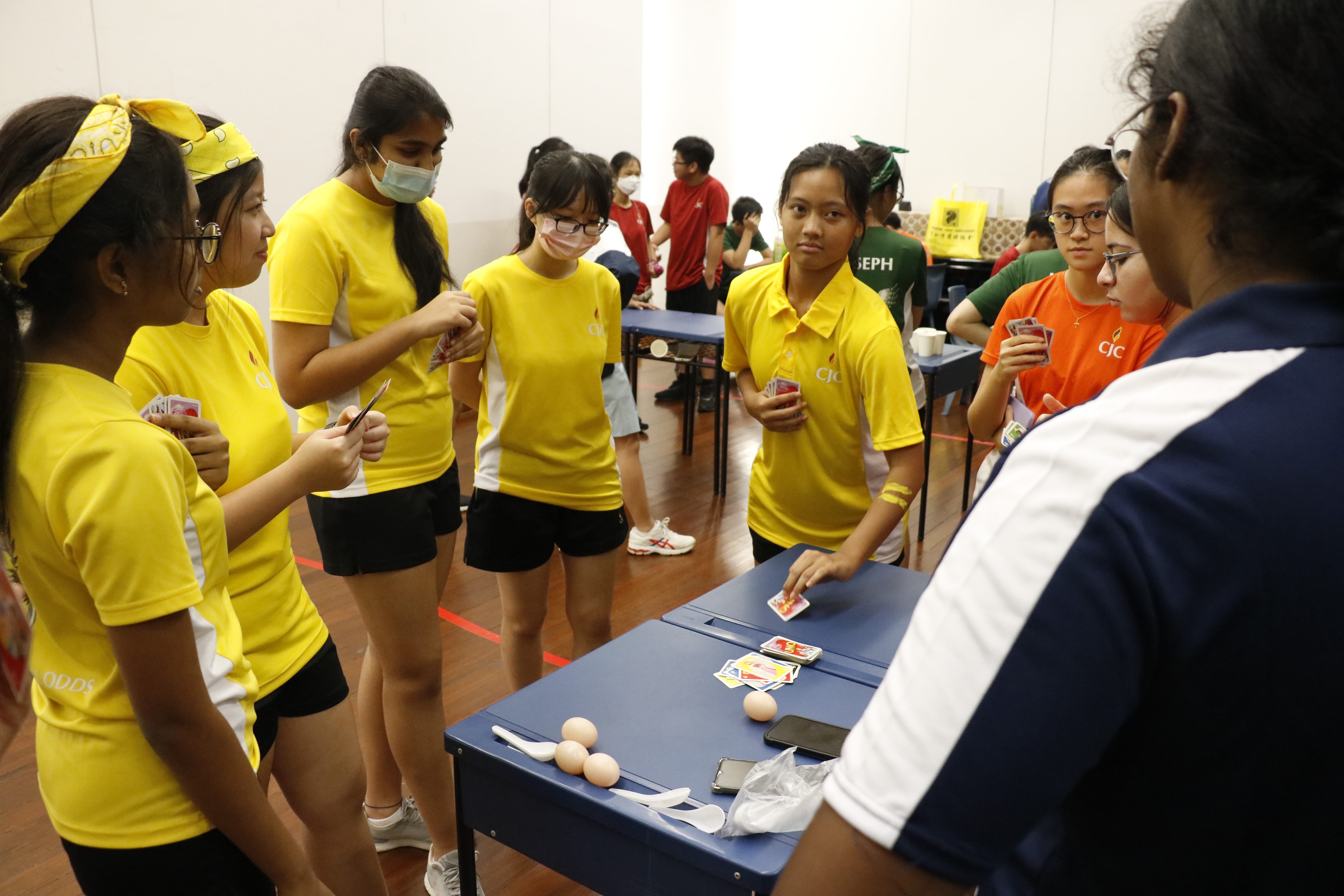 Students in yellow shirts gather around a blue table with eggs, spoons, and cards during an indoor group activity.